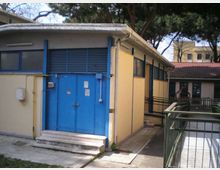 Edificio basso di servizio con facciata gialla e grandi porte metalliche blu, con piccola scalinata d’ingresso e finestre laterali. L’area esterna è pavimentata con una rampa e ringhiere, circondata da alberi e altri edifici sullo sfondo.