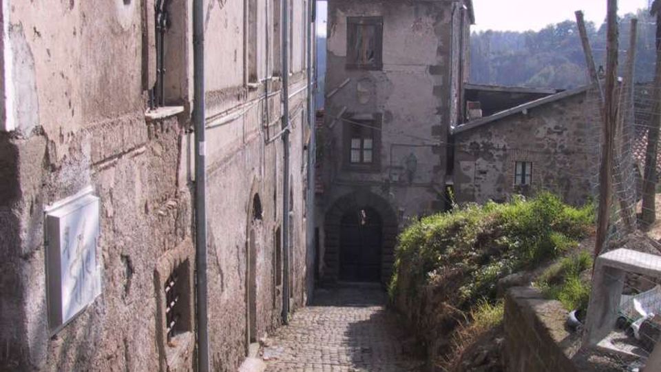 Stretta strada in acciottolato in discesa tra vecchi edifici in muratura intonacata, con finestre ad arco e lampione a parete. In fondo si vede una casa con tetto in coppi e un arco di passaggio, con vegetazione e colline sullo sfondo.
