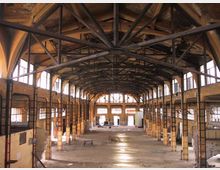 Interno di un grande capannone industriale dismesso, con struttura del tetto in travi metalliche a vista e soffitto alto. Lungo le pareti laterali ci sono file di finestre rettangolari che illuminano un ampio spazio centrale vuoto con pavimento in cemento e pilastri in muratura.