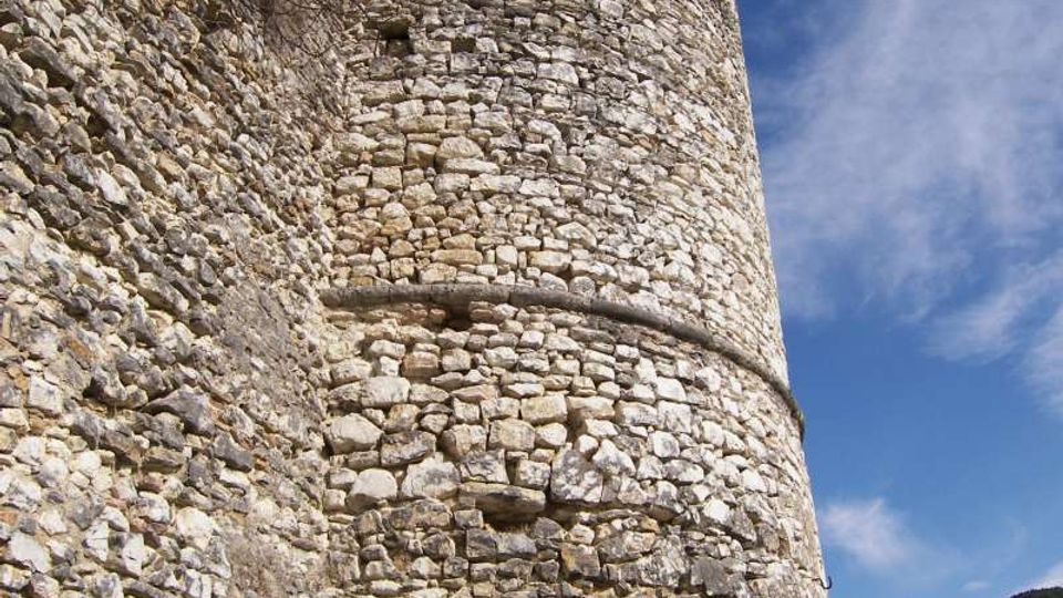 Torre cilindrica in pietra di una fortificazione, vista dal basso, con muratura irregolare e piccole aperture lungo la sommità. La struttura sorge su un pendio roccioso con vegetazione rada, sotto un cielo azzurro con nuvole sottili.