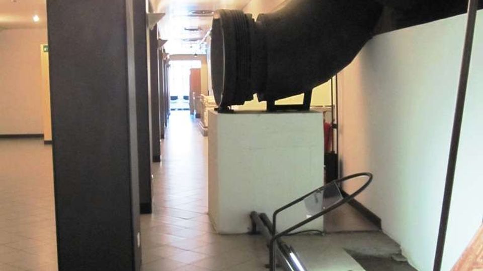 Corridoio interno di un edificio con pavimento piastrellato e soffitto basso, affiancato da colonne scure e illuminazione a parete. Sul lato destro si vedono grandi tubazioni industriali nere e una struttura metallica con pannello informativo.