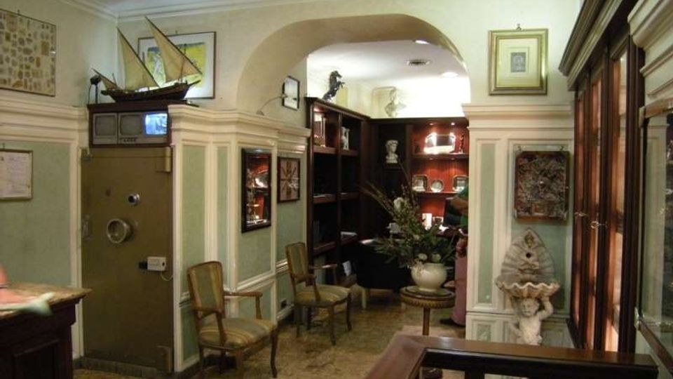 Interno di un ambiente in stile classico con pareti verde chiaro e boiserie, passaggio ad arco e pavimento in marmo. Sono visibili sedie imbottite, una cassaforte metallica, armadiature in legno scuro e scaffali con oggetti decorativi e opere incorniciate.