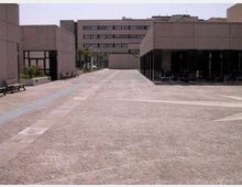 Ampio piazzale pavimentato in pietra tra edifici moderni in cemento, con panchine lungo il lato sinistro e lampioni. A destra un portico con colonne crea una zona coperta, mentre sullo sfondo si vede un edificio a più piani con finestre orizzontali.