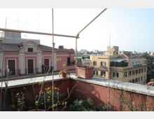 Terrazza su tetto con parapetto in muratura, vasi e piante, e struttura metallica sopraelevata. Sullo sfondo si vedono palazzi residenziali con facciate colorate e tetti in tegole, con panorama urbano in lontananza.