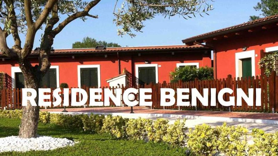 Edificio residenziale a un piano con facciata rossa, tetto in tegole e finestre con persiane verdi, circondato da una recinzione in legno. In primo piano ci sono un prato curato con aiuole e un albero, con la scritta “RESIDENCE BENIGNI” sovrapposta all’immagine.