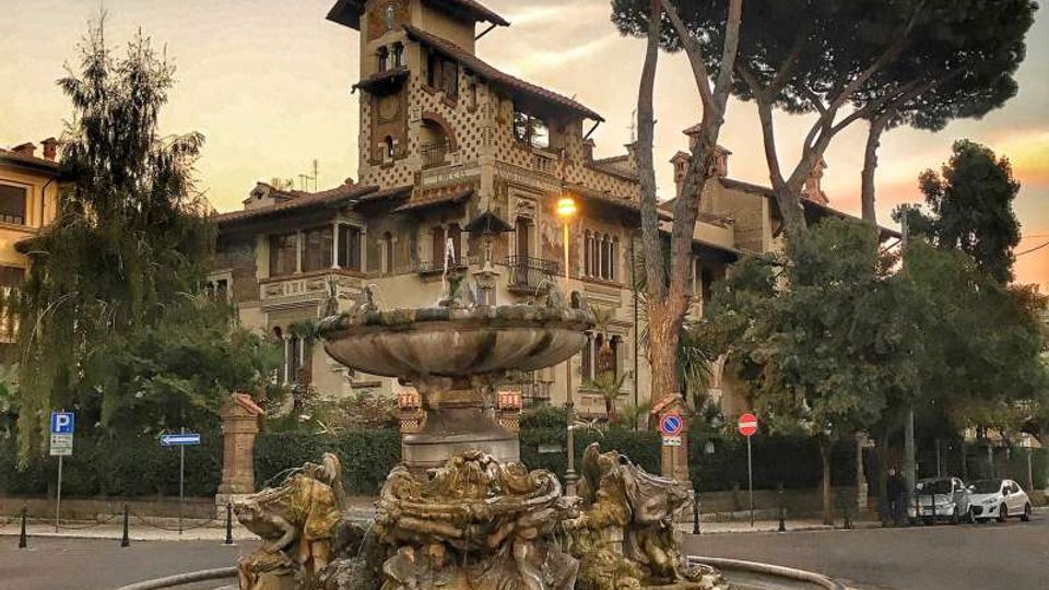 Fontana monumentale in una rotonda stradale, con grande vasca circolare e sculture in pietra alla base. Sullo sfondo si vede un edificio storico in stile liberty con torre e facciata decorata, circondato da alti pini e vegetazione urbana.