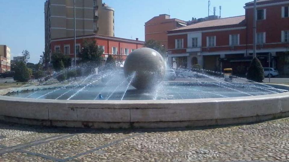 Fontana circolare in una piazza, con una grande sfera centrale da cui partono getti d’acqua verso l’esterno. Intorno si vedono pavimentazione in ciottoli e edifici urbani a più piani con facciate rosse e beige sotto un cielo sereno.