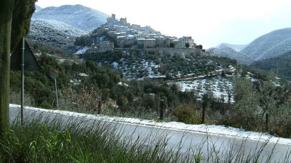 Vista panoramica di un borgo in pietra arroccato su una collina, con edifici compatti e una torre centrale, circondato da pendii e uliveti leggermente innevati. In primo piano si vede una strada asfaltata con erba alta ai bordi e segnaletica stradale, sullo sfondo colline e montagne.