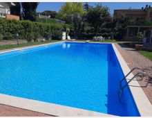 Piscina rettangolare all’aperto con acqua azzurra, bordata in pietra e circondata da pavimentazione in mattoni e prato. Sullo sfondo ci sono siepi e alberi, lettini e un tavolo con sedie vicino a edifici residenziali.