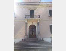 Facciata di una villa storica intonacata color crema, con scalinata in pietra che conduce a un portale ad arco incorniciato da colonne. Sopra l’ingresso c’è un balcone con ringhiera in ferro battuto e finestre con persiane scure.
