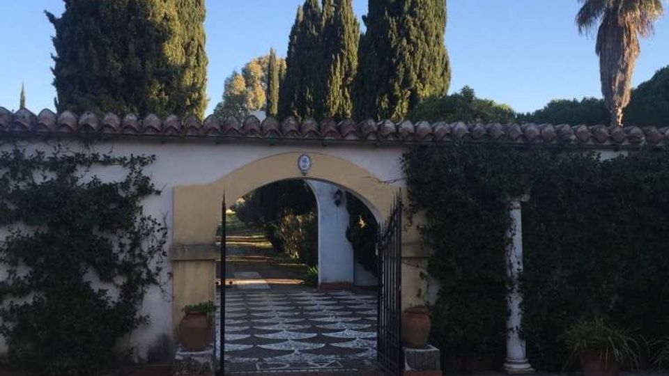 Ingresso di un giardino con cancello in ferro e arco in muratura su un muro bianco con copertura in coppi. Un vialetto pavimentato conduce all’interno, circondato da siepi e alti cipressi, con una palma sullo sfondo sotto un cielo sereno.