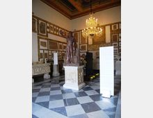 Sala museale con pavimento a scacchiera in marmo e soffitto decorato, illuminata da un grande lampadario. Al centro una statua su piedistallo, con pareti rivestite da lapidi e rilievi e pannelli informativi espositivi.