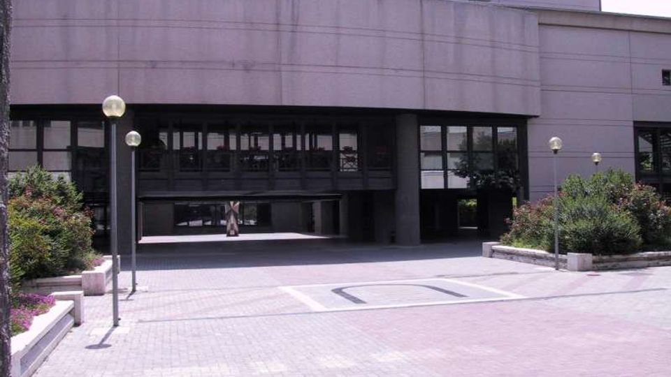 Facciata di un edificio moderno in cemento e vetro con ingresso arretrato sotto un’ampia pensilina. Davanti si apre una piazza pavimentata con lampioni sferici e aiuole rialzate con arbusti ai lati.