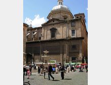 Piazza lastricata in sampietrini con una fontana centrale, circondata da dissuasori in pietra. Sullo sfondo si vede una grande chiesa in mattoni con facciata sobria e cupola ottagonale sormontata da una lanterna, sotto un cielo sereno.