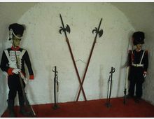 Interno di una nicchia ad arco con pareti bianche e pavimento rosso, allestita come esposizione storica con armi in asta incrociate al centro. Ai lati sono presenti due manichini in uniforme d’epoca e vari fucili e sciabole esposti su supporti.