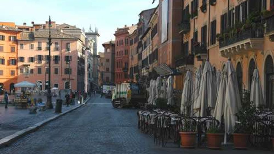 Strada lastricata in un centro storico con alti palazzi dai colori pastello e finestre con persiane. Sul lato destro si vedono tavolini all’aperto con sedie e grandi ombrelloni chiusi, affacciati su una piazza.