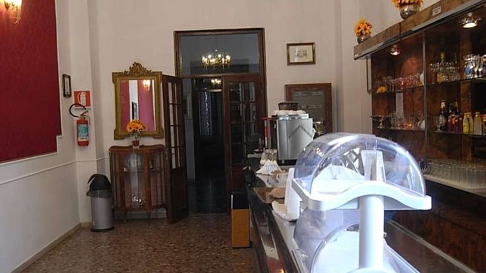 Interno di un bar stretto e allungato con pavimento in marmo, bancone in acciaio con vetrina e scaffali in legno con bicchieri e bottiglie. Lampadario classico a soffitto, pareti bianche con una sezione rosso scuro e porta a vetri sul fondo.