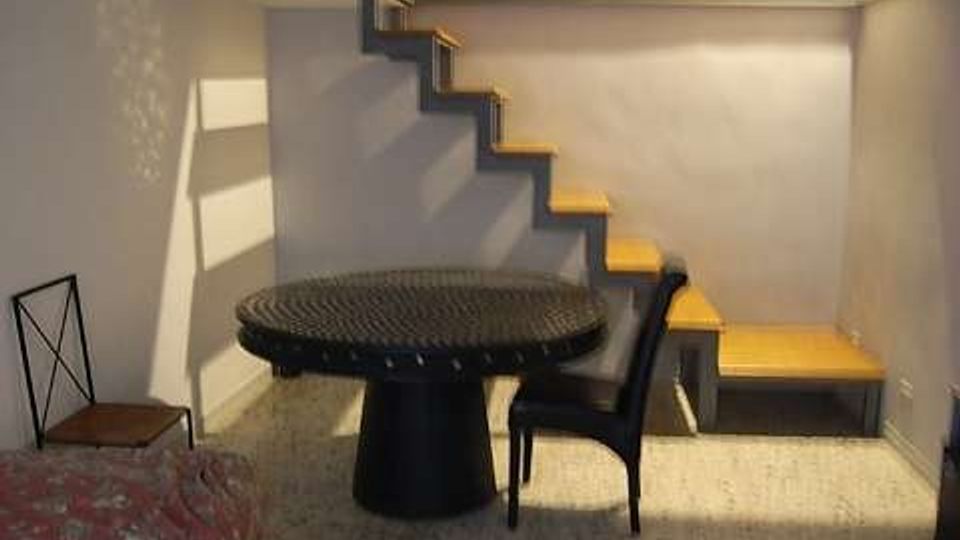 Piccolo interno seminterrato con soffitto basso, pareti bianche e pavimento in graniglia. Una scala a gradini in legno lungo la parete conduce a un soppalco, con tavolo rotondo scuro e sedie al centro e un letto singolo sul lato sinistro.