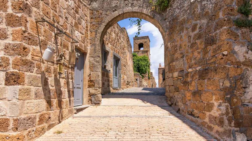 Vicolo acciottolato tra alte mura in pietra con porte grigie, attraversato da un grande arco in muratura. In fondo si vede una torre in pietra e vegetazione che cresce sulle pareti, sotto un cielo sereno.