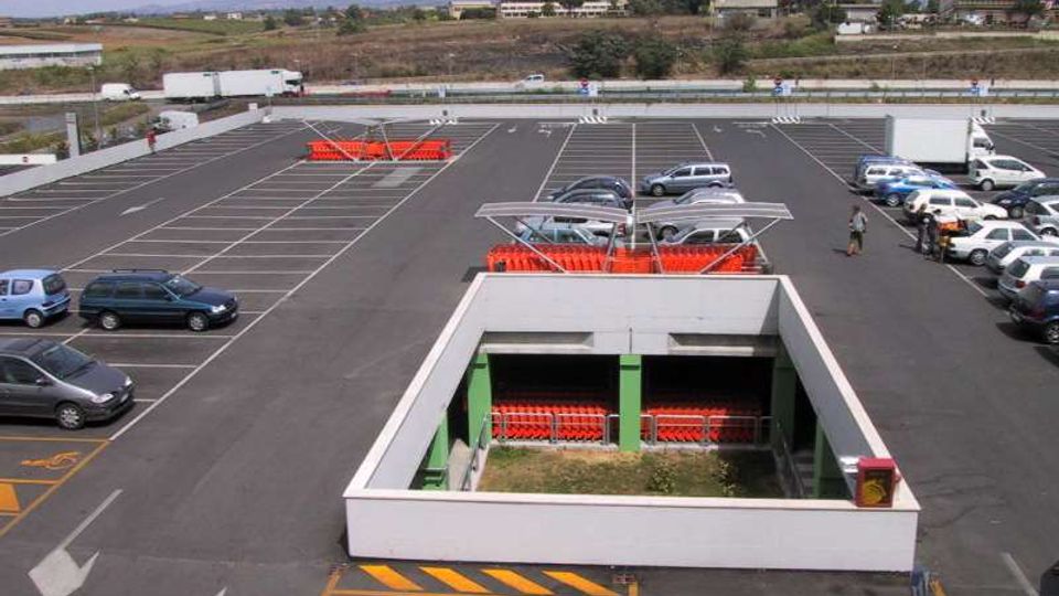 Grande parcheggio asfaltato con strisce bianche e numerosi posti auto, con alcune auto parcheggiate e aree con carrelli della spesa sotto pensiline. In primo piano si vede una struttura rettangolare incassata con pareti bianche e pilastri verdi, mentre sullo sfondo ci sono campi aperti e colline lontane.