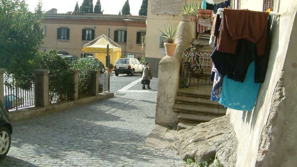 Stradina acciottolata in leggera discesa accanto a un edificio, con scala esterna e ringhiera, vasi di piante e panni stesi su un balcone. Sullo sfondo si vede una piccola piazza con auto parcheggiate, un gazebo giallo e facciate di case con alberi.