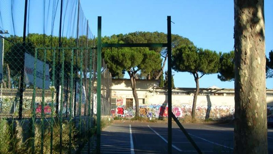 Ingresso a un campo sportivo recintato con rete metallica e pali alti, con un vialetto asfaltato che conduce a un’area di gioco segnata da linee bianche. Sullo sfondo si vedono pini e un edificio basso con graffiti, sotto un cielo sereno.