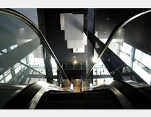 Vista dall’alto di una scala mobile in un atrio moderno con pareti scure e ampie superfici vetrate. Si vedono ringhiere metalliche lucide, finestre laterali che lasciano entrare luce naturale e pannelli geometrici sospesi sul soffitto.