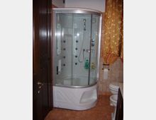 Bagno con cabina doccia angolare in vetro curvo e base bianca, dotata di soffioni e getti idromassaggio interni. Pareti e pavimento rivestiti in piastrelle color beige, con porta in legno scuro e sanitari visibili sul lato destro.