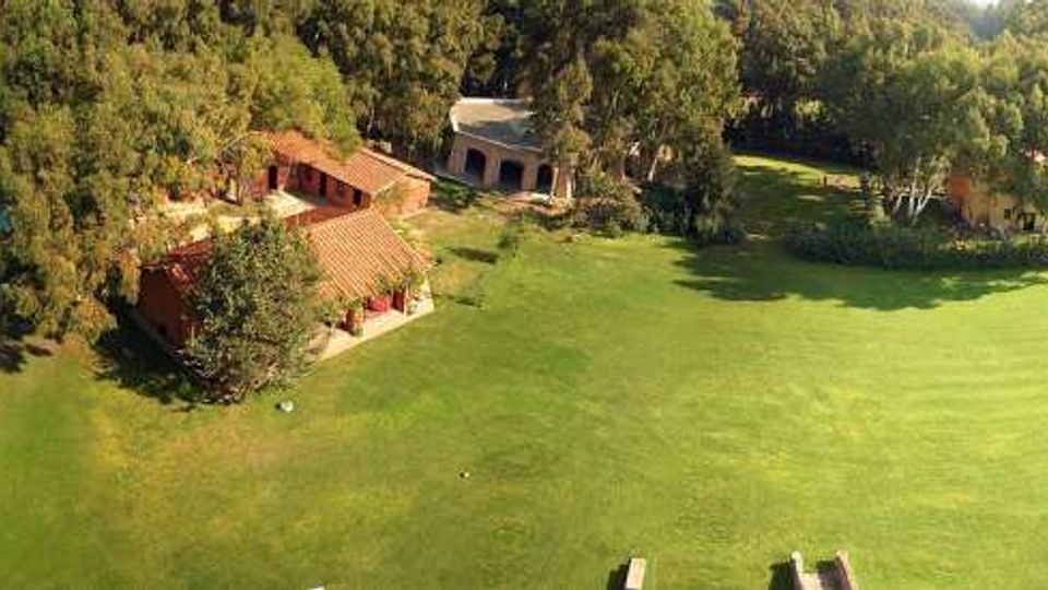 Vista aerea di una tenuta con grande prato verde circondato da alberi, edifici con tetti in terracotta e una piscina rettangolare azzurra sul lato sinistro. Sullo sfondo si vedono aree boscate e campi aperti.