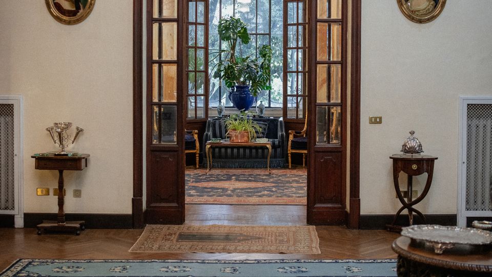 Interno di un salone elegante con pavimento in legno a spina di pesce e tappeti persiani, che conduce attraverso una grande porta ad arco in legno e vetro verso un secondo ambiente con vetrate decorative e una pianta in vaso al centro. Sulle pareti chiare sono presenti piccoli tavolini e ritratti ovali incorniciati.