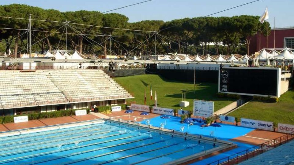 Stadio del nuoto all’aperto con piscina olimpionica a corsie, tribune in cemento e piattaforme per tuffi sul bordo. Sullo sfondo si vedono un grande tabellone elettronico, un pendio erboso, cavi di copertura e una fila di alberi alti oltre le strutture.