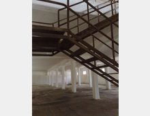 Interno di un grande capannone industriale vuoto con file di pilastri bianchi, pavimento in cemento e piccole finestre alte sulle pareti. In primo piano una scala in metallo con ringhiere attraversa lo spazio in diagonale verso un livello superiore.
