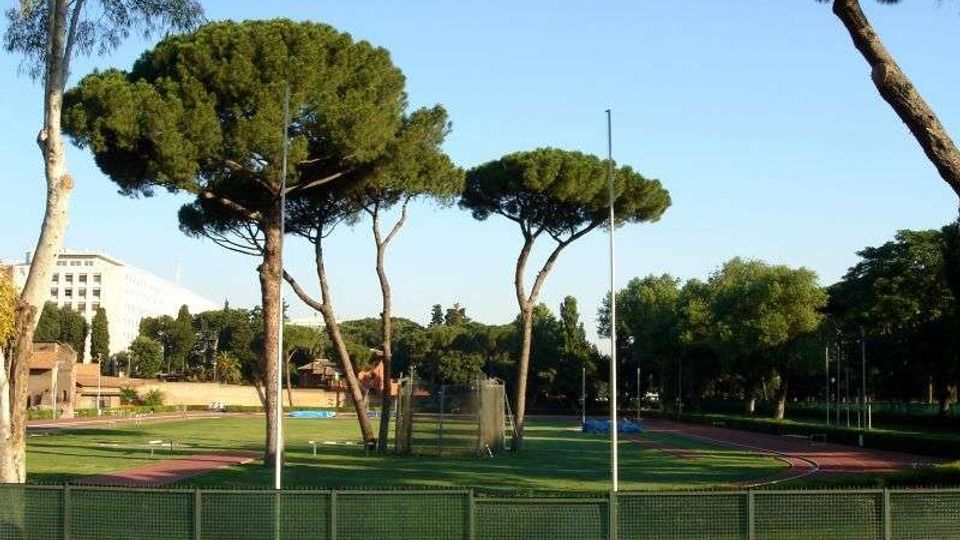 Vista di un impianto sportivo all’aperto con pista di atletica rossa che circonda un campo in erba, recintato da una rete metallica. Sullo sfondo si vedono alti pini a ombrello e altri alberi, con alcuni edifici e pali dell’illuminazione ai margini.