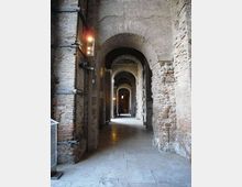 Corridoio interno in muratura antica con una sequenza di grandi archi in mattoni e pietra, pavimento in lastre di pietra e luci a parete. La prospettiva conduce verso aperture ad arco successive, creando un passaggio lungo e stretto in stile storico.