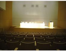 Sala conferenze moderna con file di poltroncine rivolte verso un palco rialzato, con lungo tavolo bianco per relatori e podio laterale. Pareti rivestite in legno e parete di fondo chiara con piccole aperture quadrate in alto.