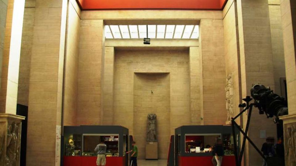 Ampio atrio interno di un museo con alte pareti in pietra chiara, soffitto a lucernario e fascia superiore rossa. Nella sala si vedono vetrine espositive rosse ai lati, una statua su piedistallo al centro e un grande oggetto scultoreo scuro sulla destra.