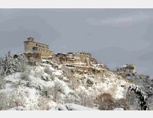Borgo in pietra arroccato su un crinale montano, con edifici storici e una struttura tipo fortezza o castello. Il pendio sottostante è coperto di neve e alberi innevati, sotto un cielo grigio invernale.
