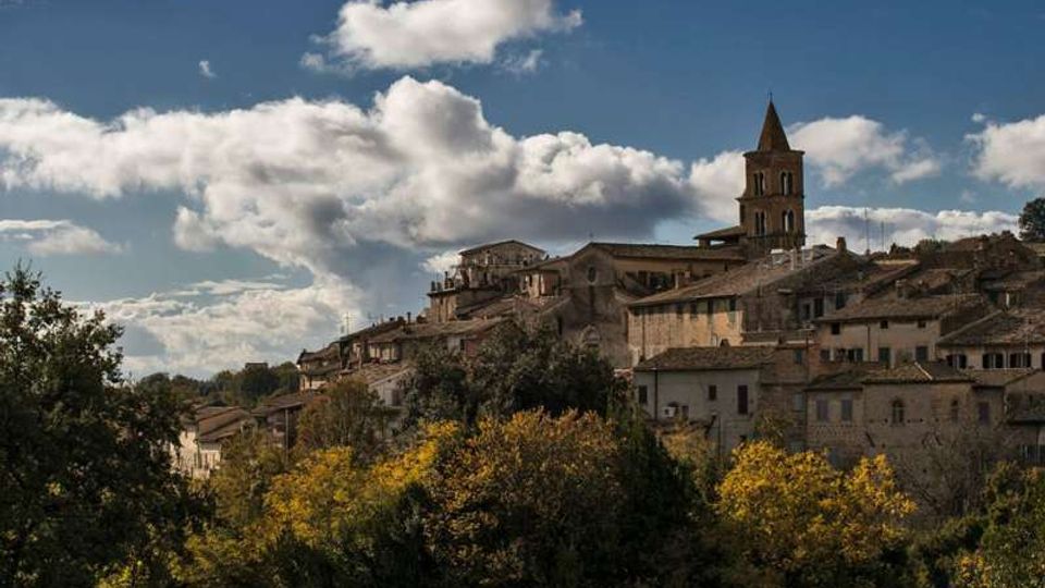 Veduta di un borgo collinare con case in pietra e tetti in coppi, dominato da un campanile con cuspide. In primo piano si vede una fascia di alberi e vegetazione, con cielo azzurro e nuvole sullo sfondo.