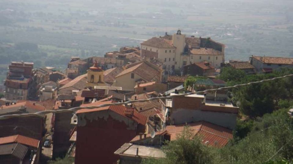 Vista panoramica di un borgo collinare con case addossate e tetti in coppi rossi, dominato da un grande edificio storico su un’altura. Sullo sfondo si estende una valle con campi e vegetazione, in lontananza leggermente velata.