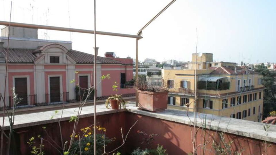 Terrazza su tetto con parapetto in muratura, vasi e piante, e struttura metallica sopraelevata. Sullo sfondo si vedono palazzi residenziali con facciate colorate e tetti in tegole, con panorama urbano in lontananza.