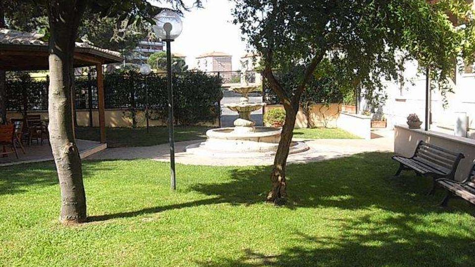 Giardino con prato verde e alberi che creano ombra, con una fontana centrale in pietra e un lampione lungo il vialetto. Ai lati si vedono una panchina vicino a un edificio e una struttura coperta tipo pergolato.