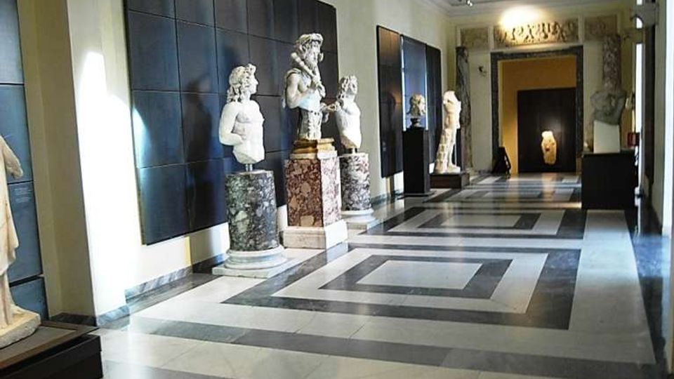 Corridoio interno di un museo o galleria con pavimento geometrico in marmo grigio e bianco e soffitto alto con illuminazione a binari. Lungo le pareti sono esposte statue e busti su piedistalli, con grandi pannelli scuri e un passaggio ad arco sullo sfondo.