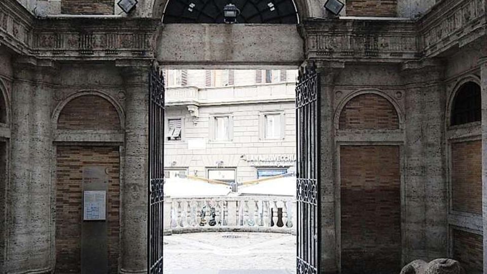 Interno di un cortile in pietra con pavimentazione a motivi geometrici e pareti con riquadri in mattoni, centrato su un grande arco con cancello in ferro battuto aperto. Oltre l’arco si vede una balaustra in pietra e la facciata di un edificio esterno.