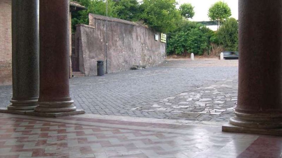 Portico con grandi colonne in pietra e pavimento a piastrelle, affacciato su una piazzetta lastricata e un’area pavimentata in pietra. Sullo sfondo si vede un lungo muro in intonaco, un lampione e vegetazione alberata lungo il perimetro.