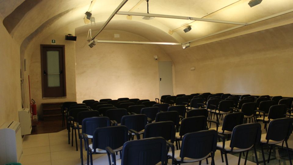 Sala conferenze sotterranea con soffitto a volta intonacato e illuminazione a binari sospesi. File di sedie nere disposte verso il fondo della stanza, con una porta laterale su un piccolo rialzo e pareti color crema.