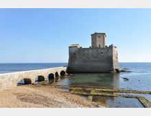 Fortezza in pietra sul mare, collegata alla riva da un ponte rialzato con più arcate. Davanti si vede una spiaggia rocciosa con pozze d’acqua e, sullo sfondo, l’orizzonte del mare sotto un cielo sereno.