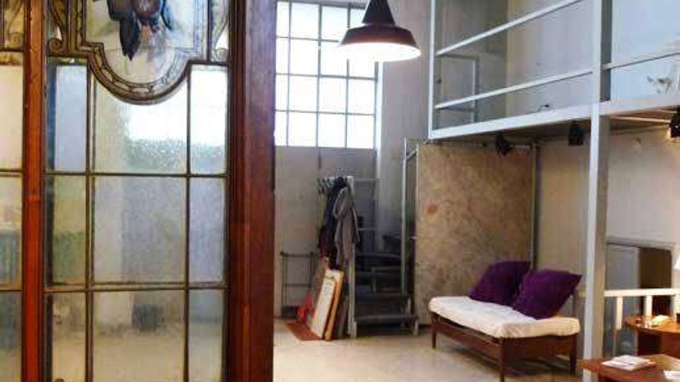 Interno di un loft con soffitti alti e grandi finestre ad arco che illuminano uno spazio open space. In primo piano si vede una porta in vetro decorato, mentre sul fondo ci sono un soppalco con ringhiera metallica, una scala, una lampada a sospensione e un divanetto.