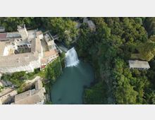 Veduta aerea di un complesso di edifici storici in pietra accanto a una cascata che si tuffa in un bacino d’acqua verde-azzurra. Intorno si estende una fitta area boschiva con sentieri e alcune strutture isolate tra gli alberi.