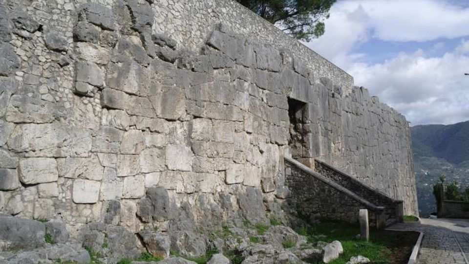 Mura esterne di una fortificazione in pietra con grandi blocchi irregolari e una piccola apertura rettangolare in alto, raggiungibile tramite una rampa inclinata. Accanto corre una strada lastricata, con erba e rocce alla base del muro e colline sullo sfondo sotto un cielo parzialmente nuvoloso.