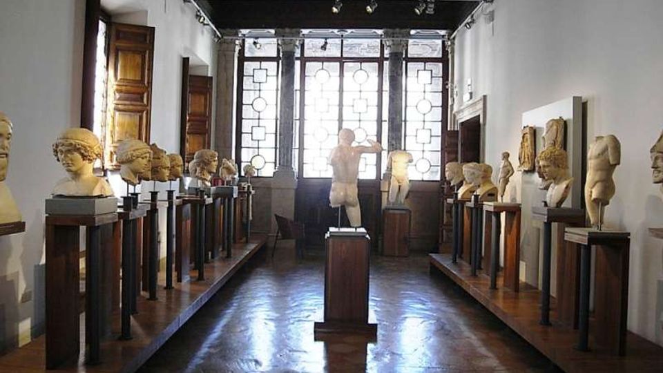 Interno di una galleria museale con pavimento in legno e file di busti e sculture su piedistalli lungo le pareti. In fondo, grandi finestre con vetri decorati illuminano la sala e una statua centrale su basamento è posizionata nell’asse del corridoio.